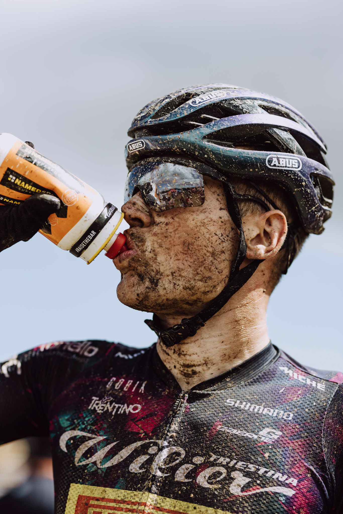 A cyclist drinking from a water bottle, his face covered in mud splatters