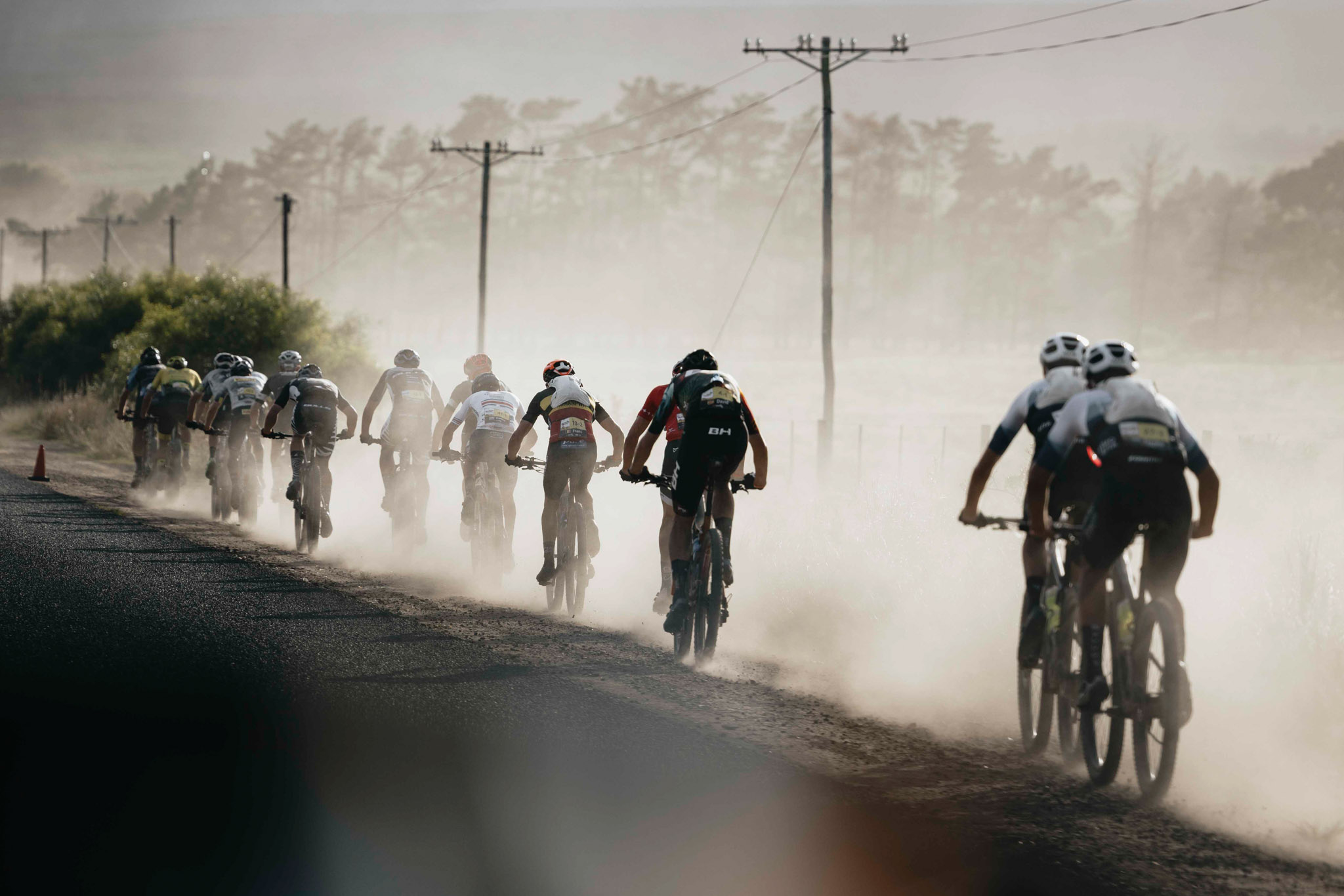 A single file of cyclists ride along a dusty road